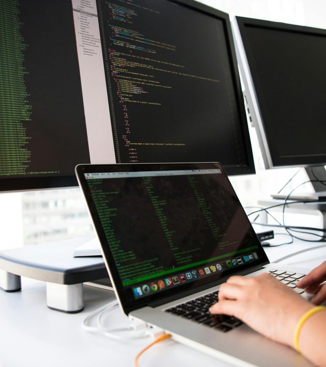 pexels-photo-1181675-1181675 A developer writes code on a laptop in front of multiple monitors in an office setting.
