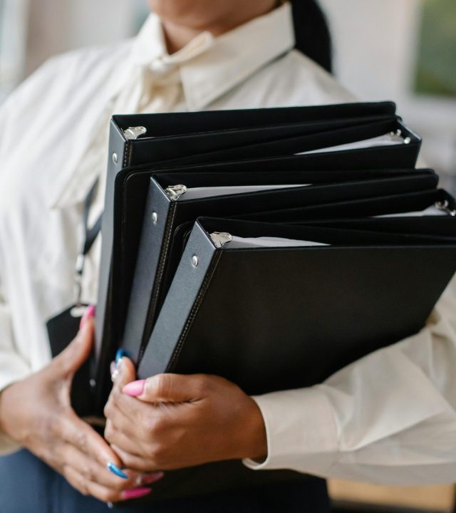 pexels-photo-5668873-5668873 Professional woman in formal attire carrying multiple black folders indoors.